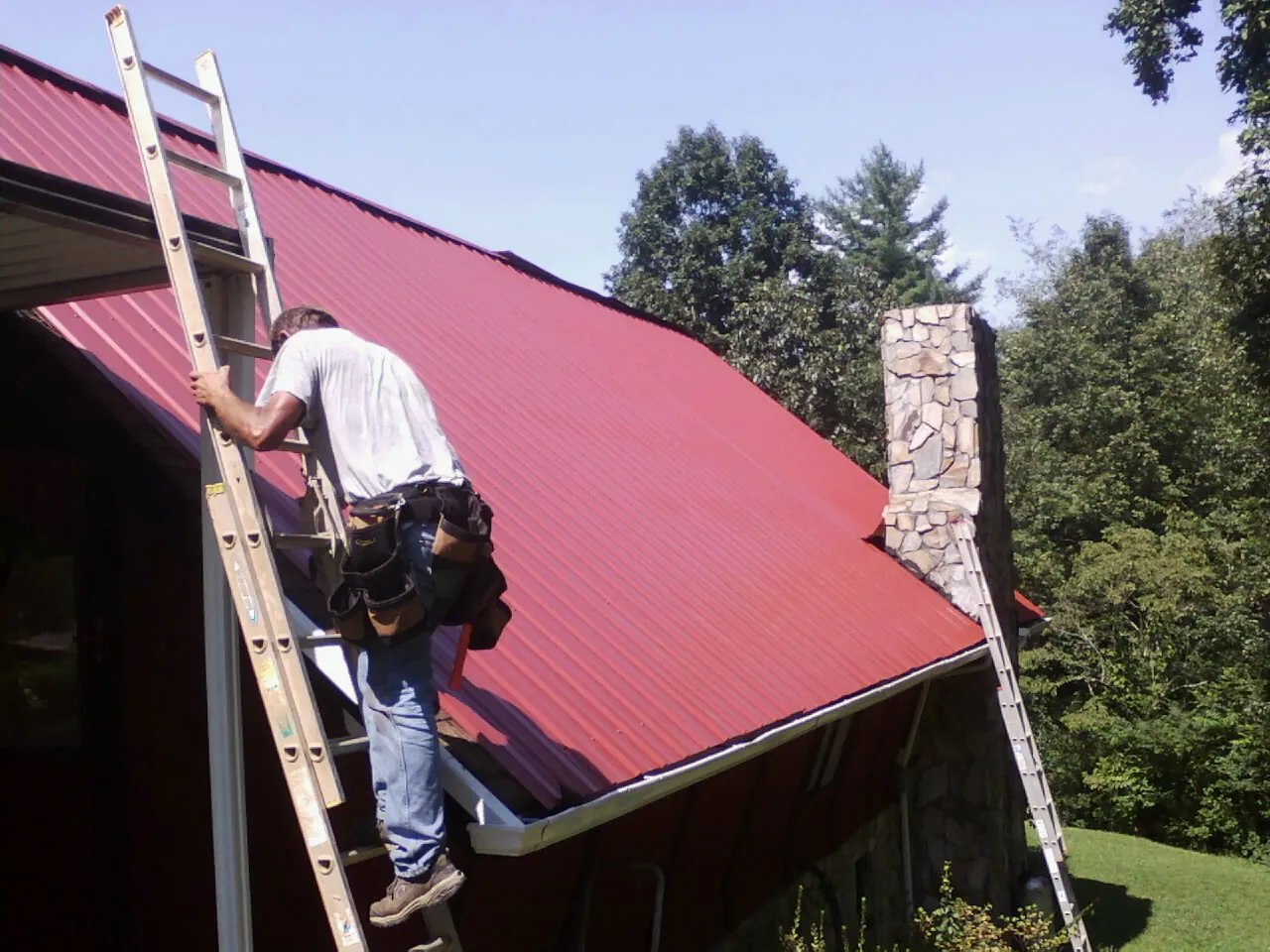 Roofer on a ladder beside a red metal roof and stone chimney — Soffit & Fascia Repair by Roofing Brothers in Washington Depot